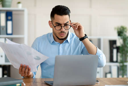 Stressed Arab employee with laptop and documents having problem with work, making mistake in business project at officeの写真素材