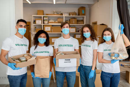 Group portrait of young diverse volunteers carrying donation boxes, posing indoors and wearing medical masks and glovesの写真素材
