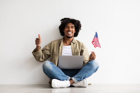 Happy Young Black Guy Sitting On Floor With Laptop And American Flagの写真素材