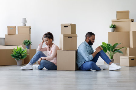 Unhappy young black guy and lady are tired and sitting on floor in apartment with boxes