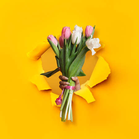 Black woman holding bouquet of fresh bright tulips through hole in ripped orange paper, closeupの写真素材