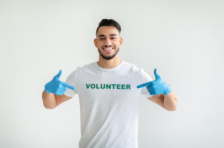 Young arab guy showing volunteer t-shirt and smiling at camera, being proud and happy of working at charity centerの写真素材