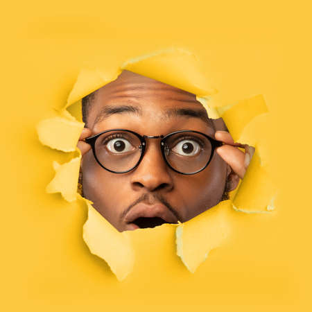 Amazed shocked millennial black male in glasses open mouth looks through hole in yellow paper,...