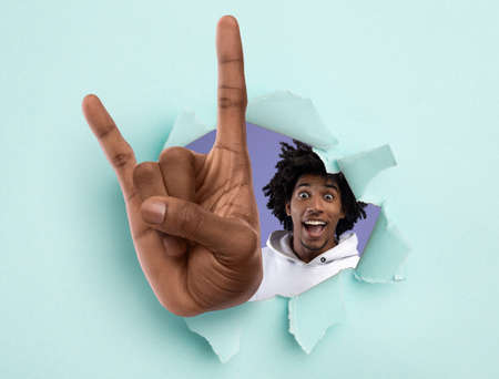 Excited black teenager showing huge rock gesture through hole in light blue ripped paperの写真素材