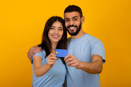 Easy contactless payments. Excited arab couple with credit card hugging and smiling at camera over yellow backgroundの写真素材
