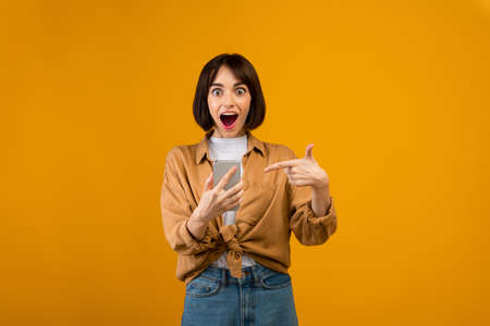Emotional lady pointing at smartphone in excitement, winning lottery or receiving amazing news, yellow backgroundの写真素材
