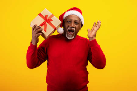 Excited senior black man in red sweater and Santa Claus hat holding gift box, shouting HO HO HO, offering big Xmas saleの写真素材
