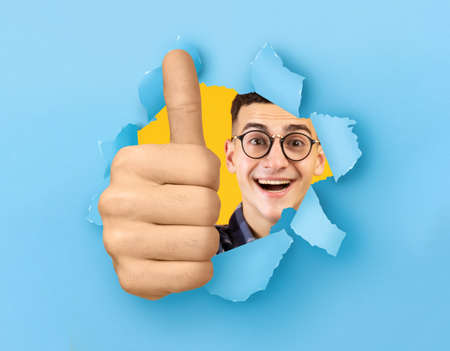 Happy young Caucasian man showing thumbs up, doing approval gesture through hole in ripped blue paperの写真素材