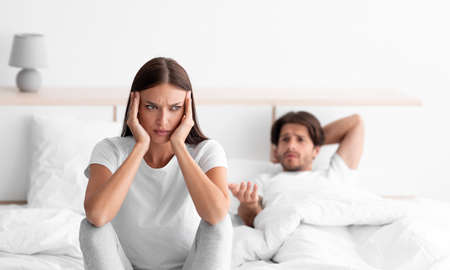 Upset unhappy caucasian young wife ignores husband, guy lies in bed in white bedroom interior,...