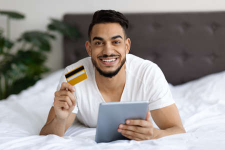Online Shopping. Arab Man With Digital Tablet And Credit Card In Bed