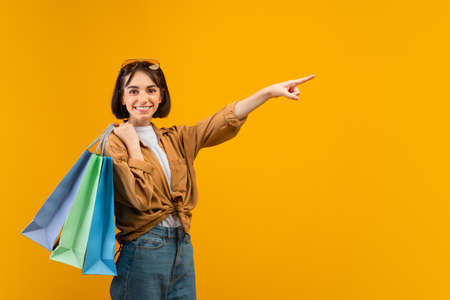 Positive lady holding shopper bags, pointing aside at free space on yellow studio background and smiling at cameraの写真素材