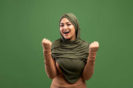 Great bonus. Joyful arab lady in headscarf celebrating success and shaking fists over dark green backgroundの写真素材