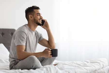 Arab Man Talking On Cellphone And Drinking Coffee While Relaxing In Bedの写真素材