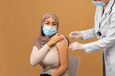 Doctor making injection of anti-covid vaccine to muslim woman in face mask and hijab, sitting over brown backgroundの写真素材
