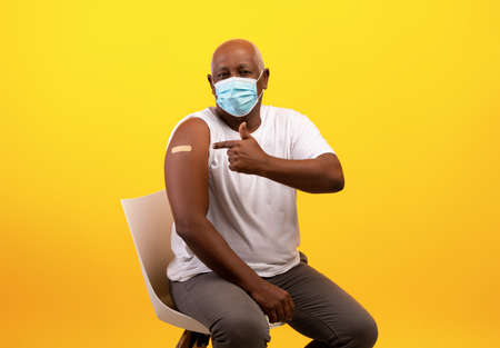 Senior black male in face mask pointing at band aid after getting coronavirus vaccine shot on orange backgroundの写真素材