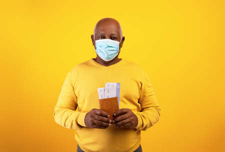 Senior black man in medical mask holding plane tickets and international passport on orange studio backgroundの写真素材