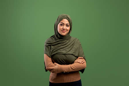 Muslim beauty concept. Portrait of young arab woman in hijab posing with folded arms over dark green backgroundの写真素材