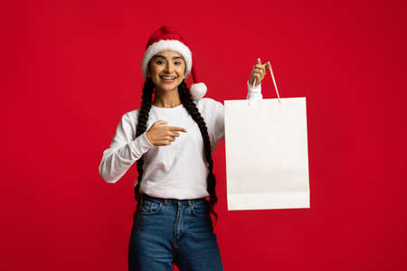 Happy Arab Woman In Santa Hat Pointing At Blank Paper Shopping Bag