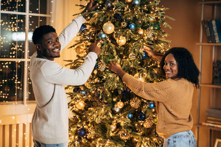 Happy young black couple decorating beautiful Christmas tree together at homeの写真素材