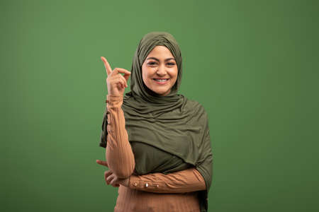 Positive islamic lady in hijab having idea, pointing finger up, having inspirational moment over olive backgroundの写真素材