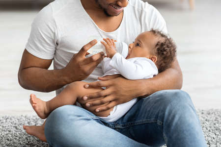 Adorable Black Baby Drinking Water Form Bottle While Lying On Fathers Lapの写真素材