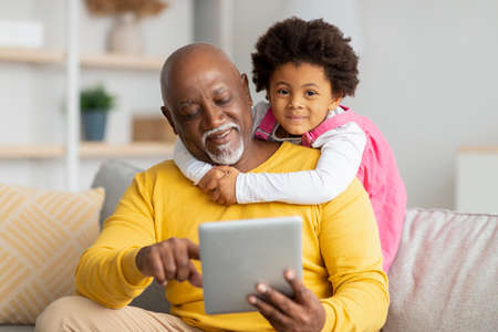 Happy african american preschooler girl hugs mature man surfing in internet or playing online game...