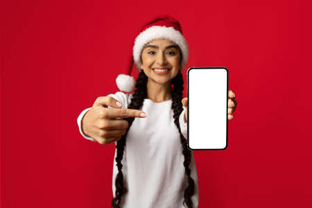 Christmas Sale. Cheerful Arab Lady In Santa Hat Pointing At Blank Smartphone