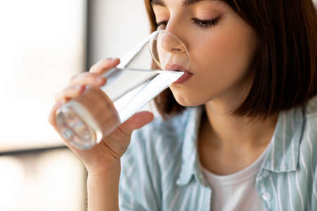 Healthy lifestyle, diet and water balance. Young lady drinking water from glass in kitchen interior,...