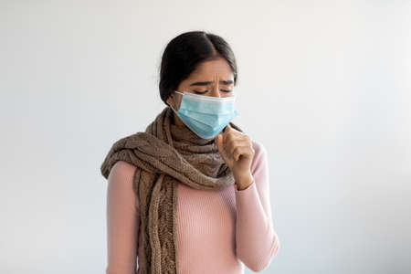 Upset despaired young indian lady in protective mask and scarf coughs, feeling bad and suffers from illnessの写真素材