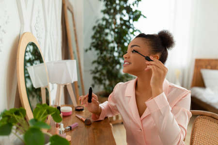 Young african american lady looking at mirror and applying mascara, sitting in bedroom interior, copy spaceの写真素材