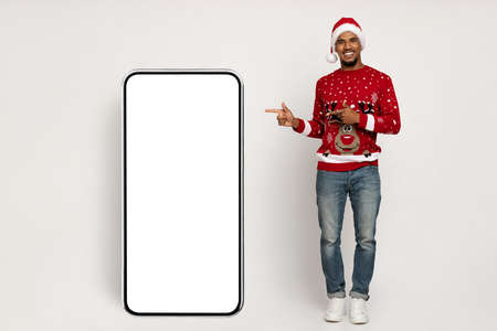 Cheerful black man in santa hat pointing at big smartphone