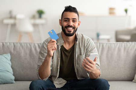 Ordering goods online. Happy young Arab guy with credit card and smartphone shopping on web at homeの写真素材