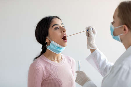 European doctor doing covid-19 pcr test for young indian woman on gray wall background in clinicの写真素材