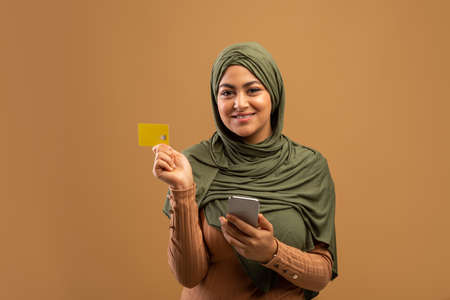Banking, purchasing concept. Cheerful muslim woman holding smartphone and credit card, buying goods in web storeの写真素材
