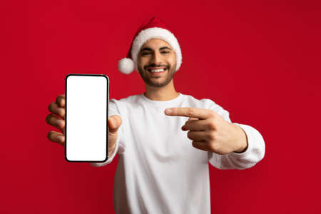 Online Offer. Cheerful Arab Guy In Santa Hat Pointing At Blank Smartphoneの写真素材