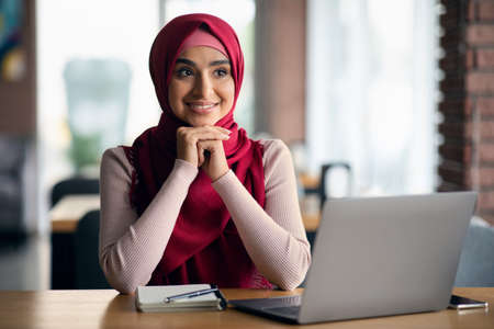 Excited young woman in hijab working on laptop, cafe interiorの写真素材