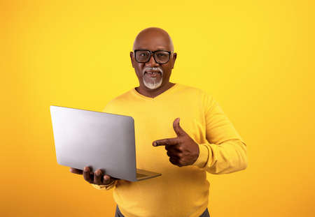 Senior African American man in glasses pointing at laptop computer on orange studio backgroundの写真素材