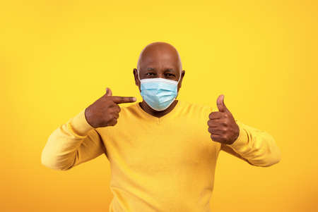 Senior black man pointing at mask, showing thumb up, recommending facial protection during...