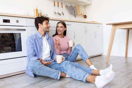 Smiling young european lady and man with cups, drink coffee, sitting on floor, talking in spare timeの写真素材