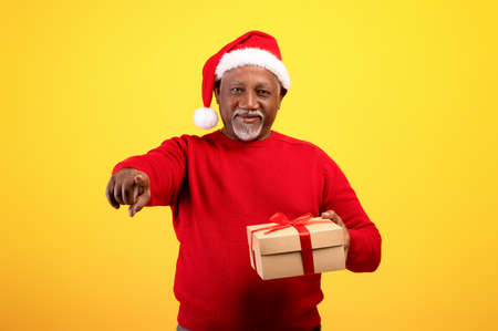 Portrait of happy senior black man in Santa Claus hat holding Christmas gift, pointing at you on orange backgroundの写真素材