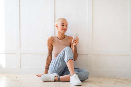 Young stylish lady sitting on floor with smartphone, working remotely or video chatting against white wall, free spaceの写真素材