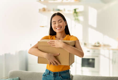 Overjoyed asian lady hugging carton parcel, receiving long awaited delivery, getting online order,...