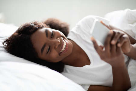 Cheerful african american woman using cellphone in bedの写真素材