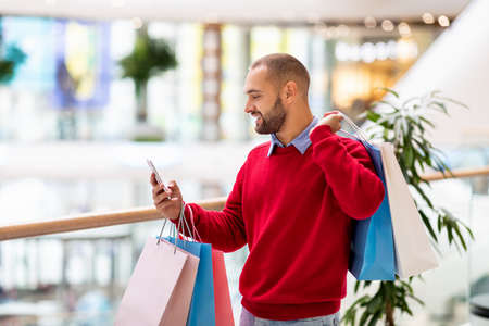 Happy young man with colorful gift bags using cellphone at mall, checking shopping list, having video call, copy spaceの写真素材