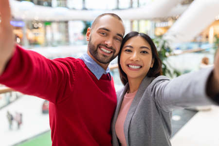 Loving young diverse couple smiling, taking selfie at huge shopping centreの写真素材
