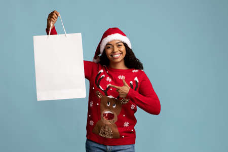 Cheerful lady in Santa hat pointing at white shopping bag