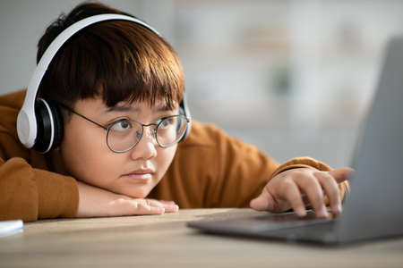 Closeup of bored asian kid wearing glasses studying online