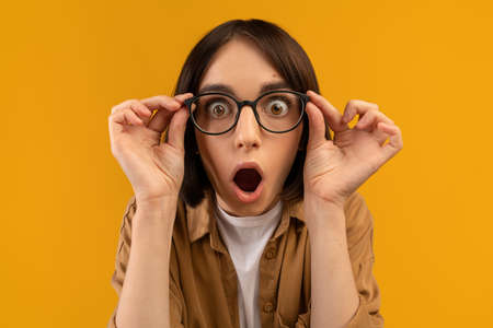 Unbelievable. Shocked caucasian lady touching glasses and looking at camera in shock, posing over yellow backgroundの写真素材