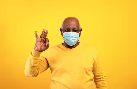 Senior black man in medical face mask showing okay gesture over orange studio background. Stay safe conceptの写真素材