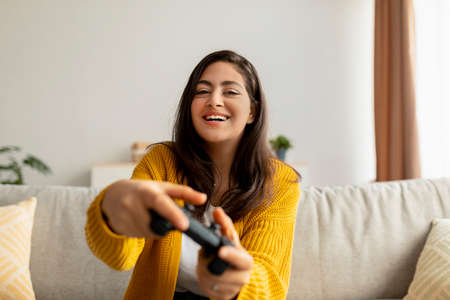 Excited middle eastern lady playing video game with joystick at home, sitting on sofa and smiling,...